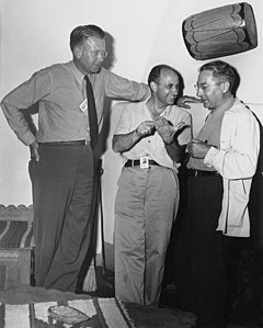 Three men talking. The one on the left is wearing a tie and leans against a wall. He stands with his head and shoulders visibly above the other two's heads. The one in the center is smiling, and wearing an open-necked shirt. The one on the right wears a shirt and lab coat. All three have photo ID passes.